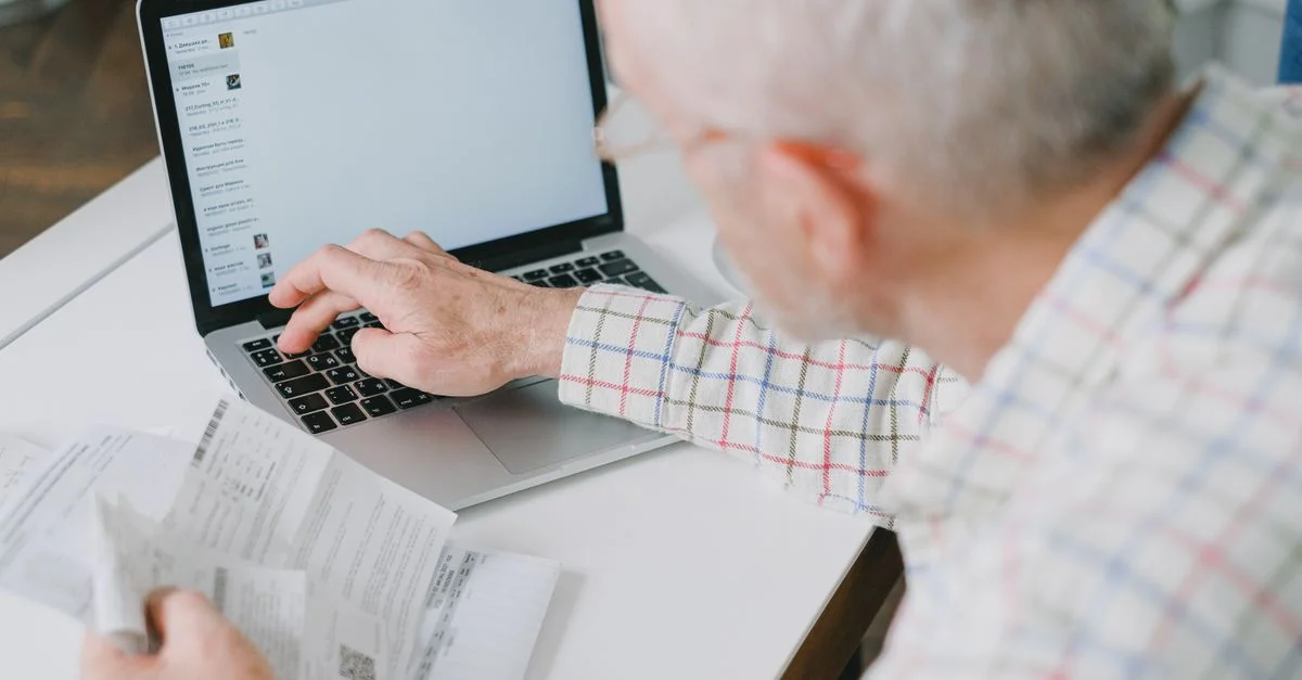 Un homme âgé à son bureau gérant ses finances en ligne avec des documents et un portable.