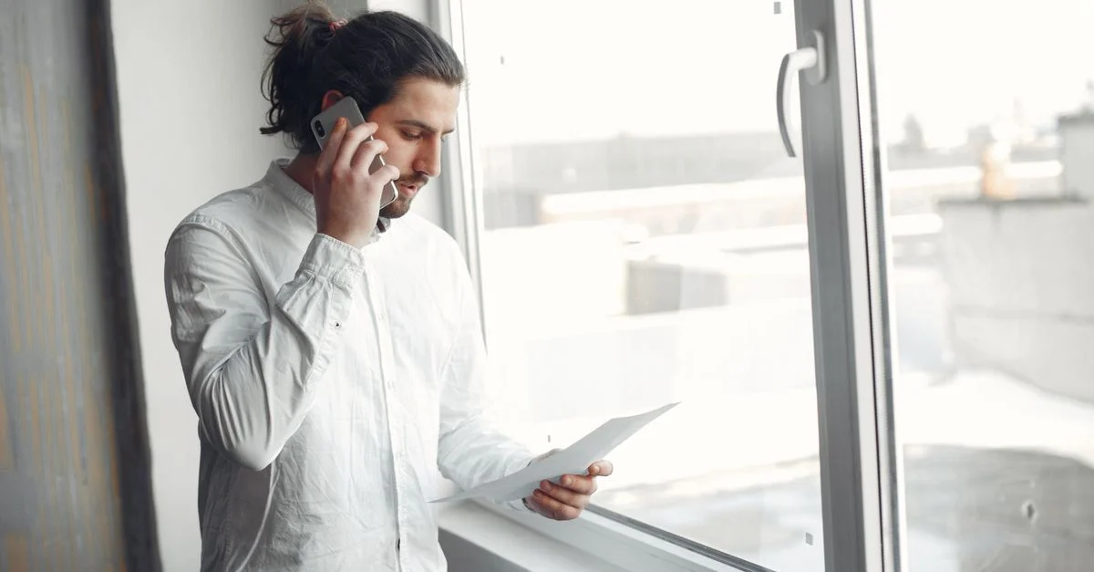 Jeune homme en chemise blanche, au téléphone tenant un document, debout près d'une grande fenêtre.