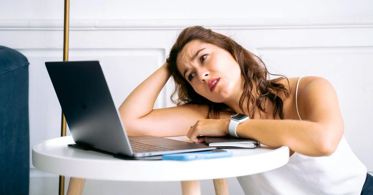 Une femme frustrée est assise à un bureau, la main sur la tête, fixant un ordinateur portable ouvert.