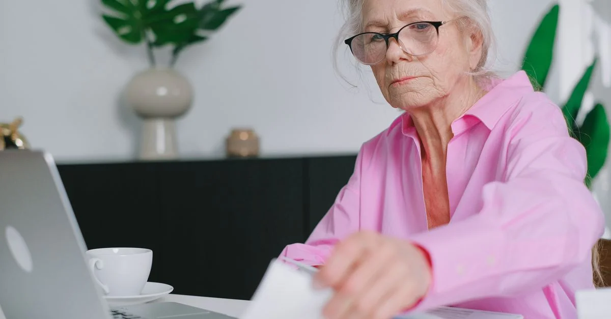 Femme âgée en chemise rose travaillant sur un ordinateur portable à la maison, concentrée et s'engageant avec des documents.